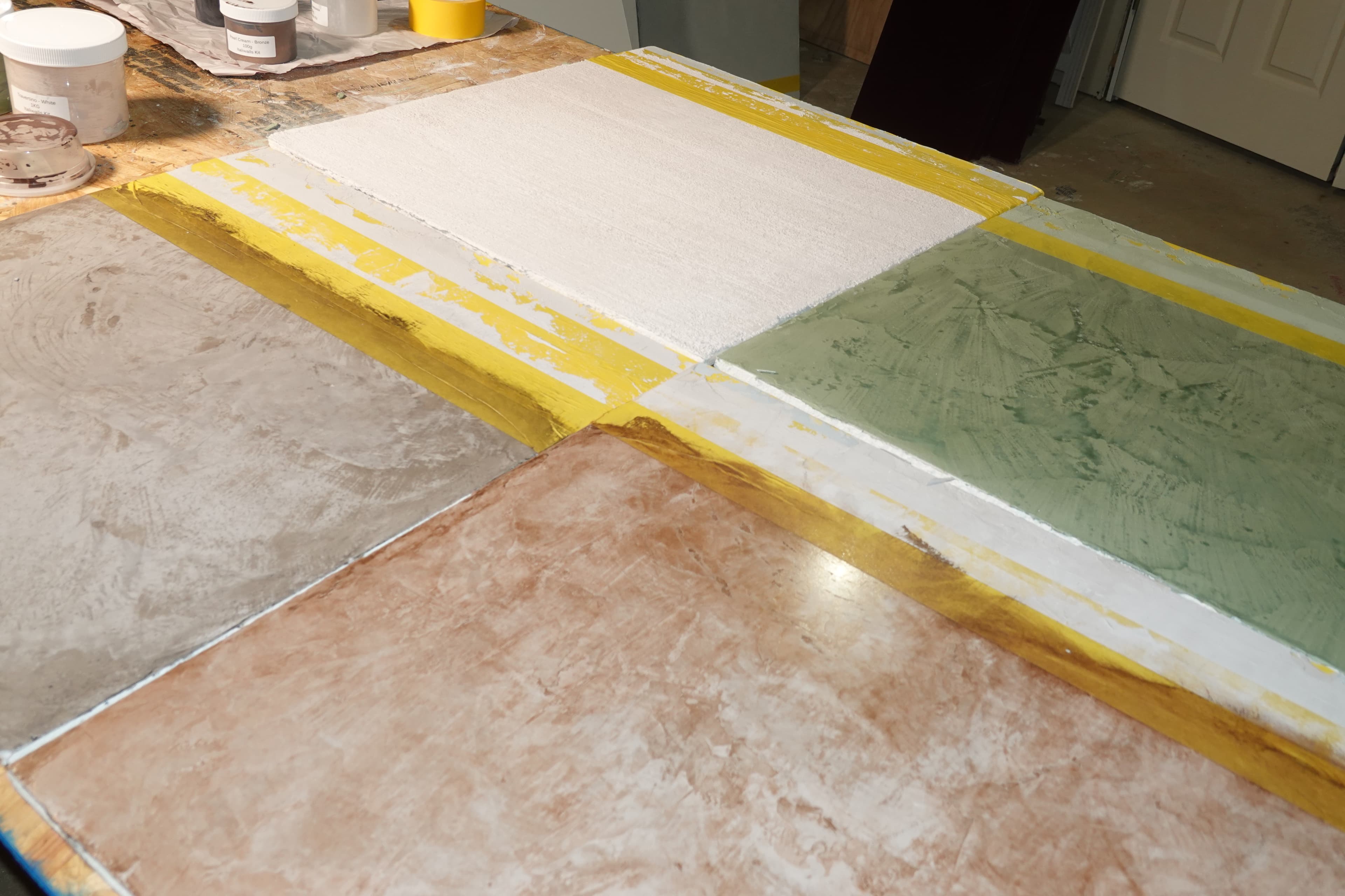 Four taped lime-plaster sample boards laid out together on the studio bench, in gray, white, sage, and terracotta, showing the range of color and texture developed for a project.